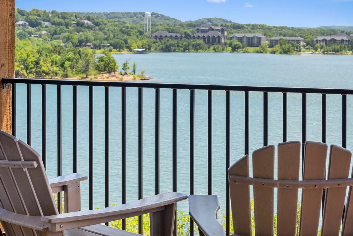 The image showcases two inviting wooden chairs positioned on a deck overlooking the tranquil expanse of Table Rock Lake, with rolling green hills and a few distant buildings enhancing the feeling of peaceful seclusion. The scene embodies relaxation, with ample natural light and a sweeping view that invites guests to sit, unwind, and take in the sunset or enjoy stargazing at night.
