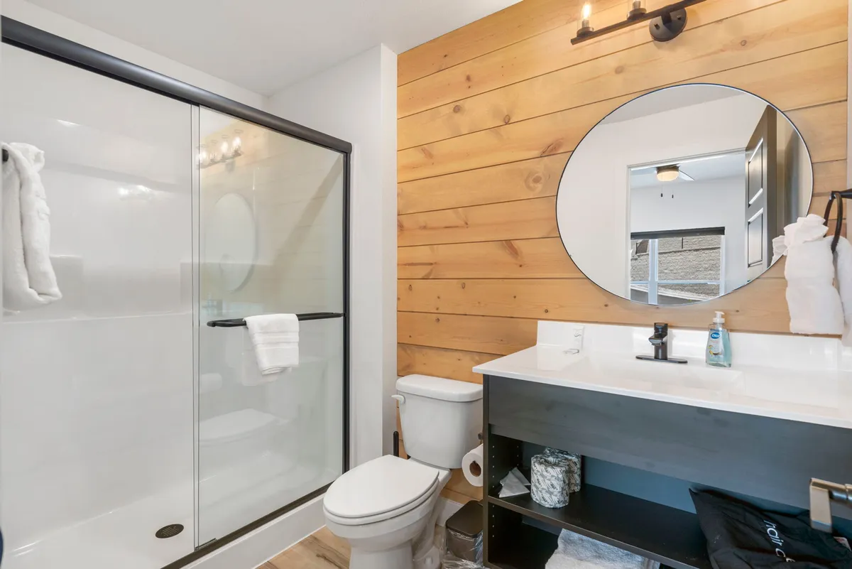 A refreshing, modern bathroom with a glass shower enclosure, wood-paneled wall, round mirror above a sleek sink, and a calming atmosphere. Designed for relaxation and personal care, the space completes the sanctuary experience.