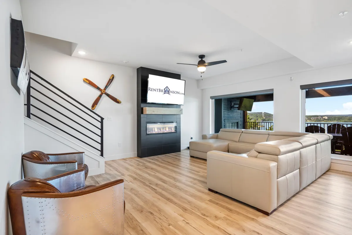A comfortable living area featuring a leather sectional sofa, a modern fireplace, a TV screen, and a wooden staircase. The design is contemporary and cozy, with windows that let in light and connect the space to the outdoors.