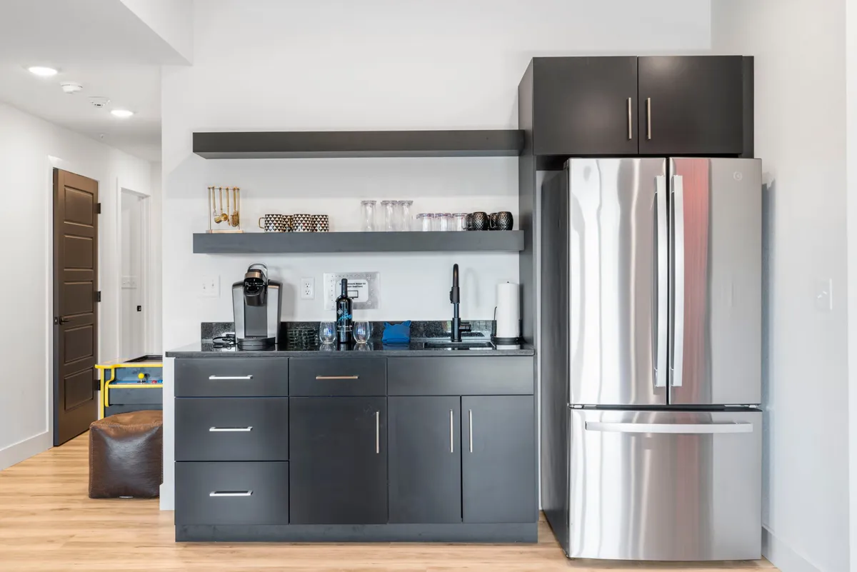 A modern kitchenette and bar area with a gleaming stainless steel refrigerator, black cabinetry, and a countertop with a sink and coffee maker. It feels sleek yet functional, ideal for preparing snacks and drinks for a group.