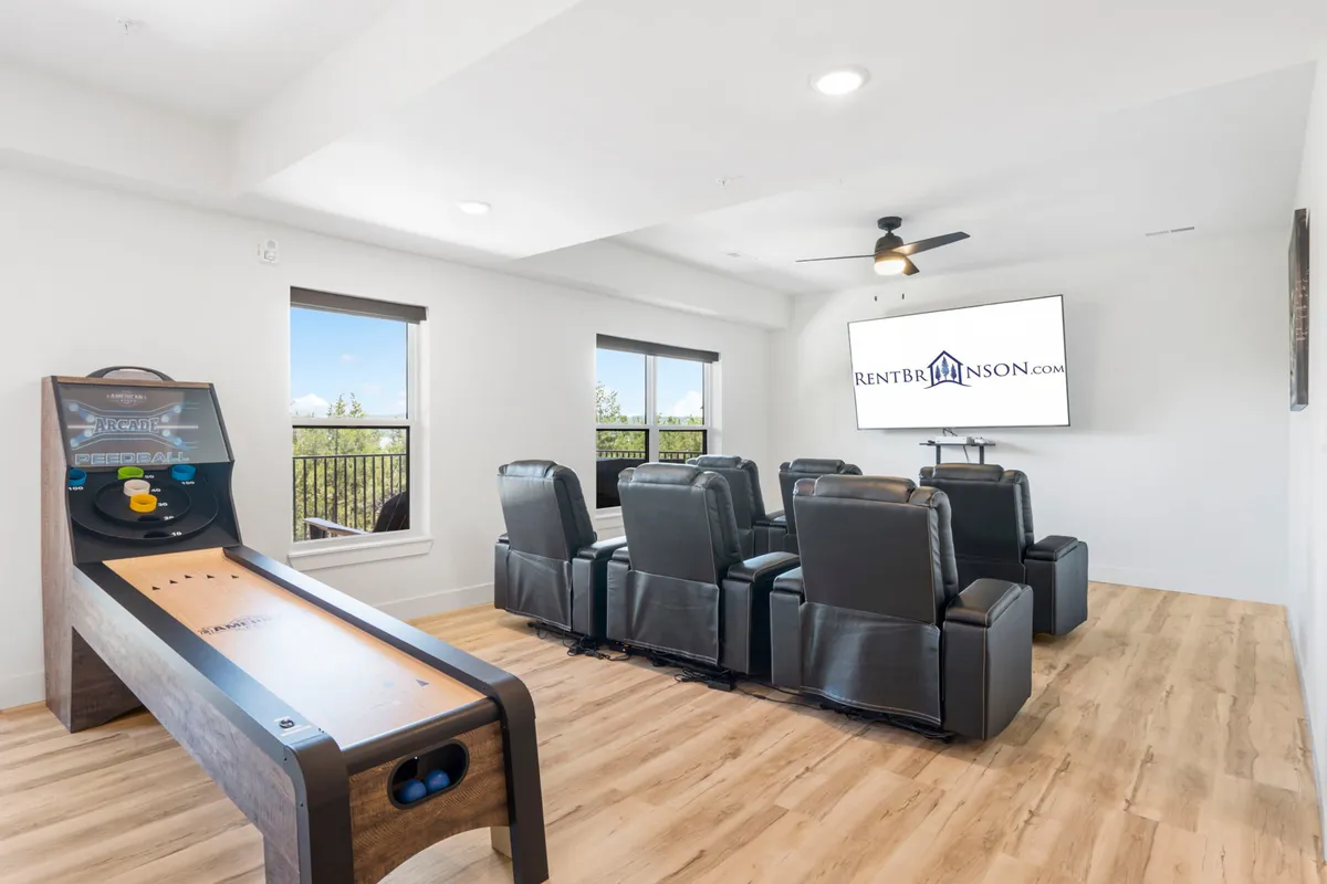 A spacious entertainment area with comfortable seating around a big screen TV, a shuffleboard table for games, and large windows providing brightness and openness. The space invites socializing, watching sports or movies, and casual group fun.