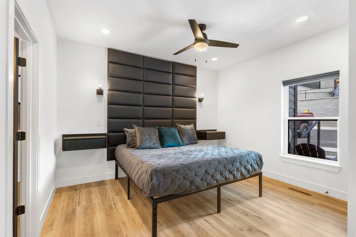 A bright and contemporary bedroom featuring a cozy bed with decorative pillows, a sleek black tufted headboard, and a ceiling fan above. Soft natural light pours in, illuminating the serene, uncluttered space that invites relaxation and restful sleep.