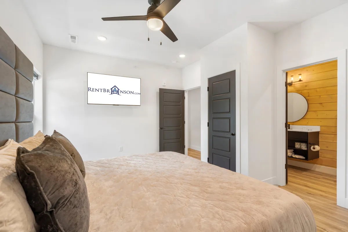 A stylish, relaxing bedroom featuring a plush bed adorned with soft pillows, a wall-mounted TV, a sleek ceiling fan, and dark wood double doors leading into a modern ensuite bathroom. The space exudes comfort and privacy.