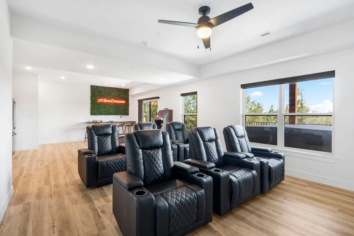 A luxurious, purpose-designed movie theater room featuring plush reclining chairs arranged in rows, a glowing neon sign, and large windows letting in natural light. The setting is stylish and dedicated to immersive movie-watching or special group celebrations.