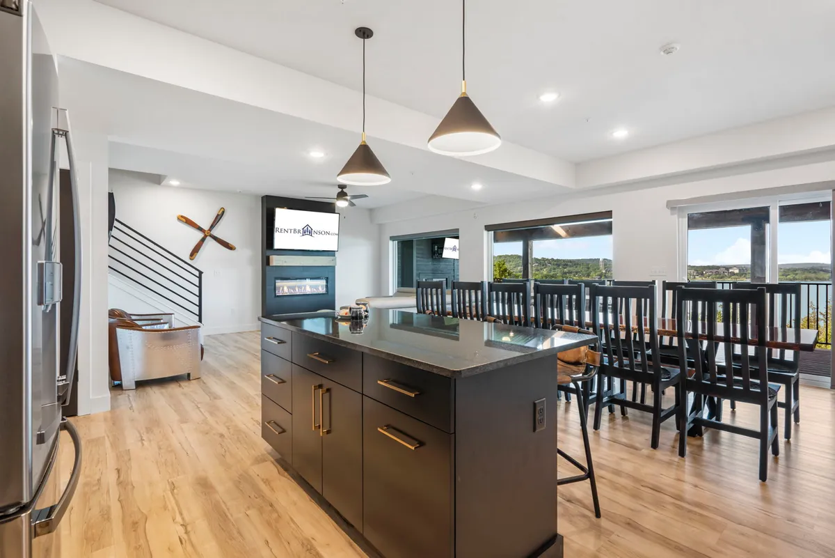 This image showcases an open-concept layout with a kitchen island, dining table, large windows, and contemporary lighting fixtures. The space is bathed in natural light, and the arrangement encourages easy movement between cooking, dining, and relaxing areas.
