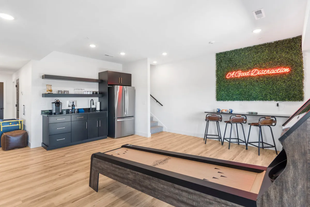 A playful entertainment nook featuring a shuffleboard table, neon sign, bar seating, and a kitchenette, evoking a lively game night or celebration. The area is inviting and set up for both play and refreshment.